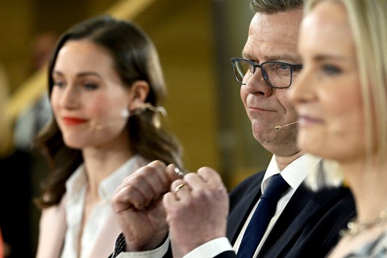 A man in a suit and glasses gestures with his fists. He is flanked by a dark-haired woman on his right and a woman with blond hair on his left.