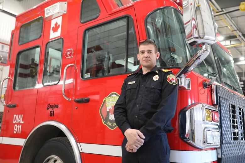 A white man, wearing his black firefighter uniform, stands in front of a big red firetruck.