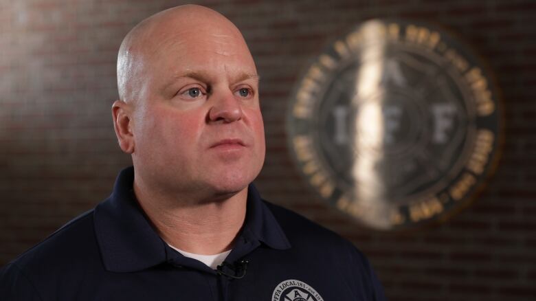 A white bald man, with a clean-shaven face, is wearing a navy blue polo shirt. His organizations logo is on the left breast, in grey. He is standing in front of a brick wall, with a silver sign showing his organizations logo.