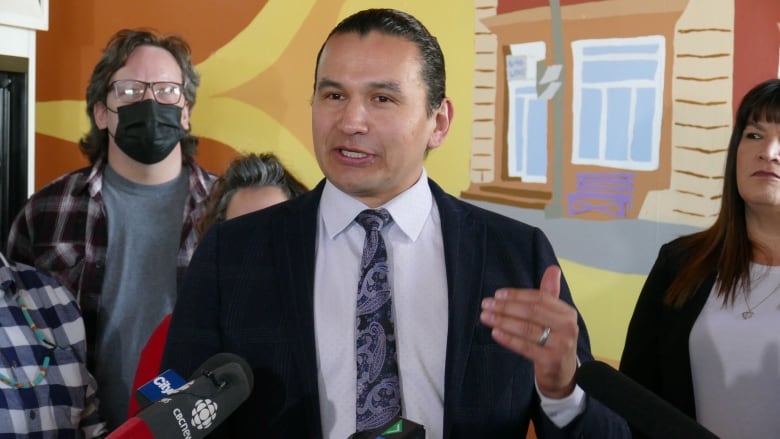 A man in a dark suit with a light shirt and tie speaks into microphones at a news conference.