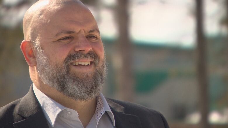 A man with a grey beard in front of a blurry outdoor background