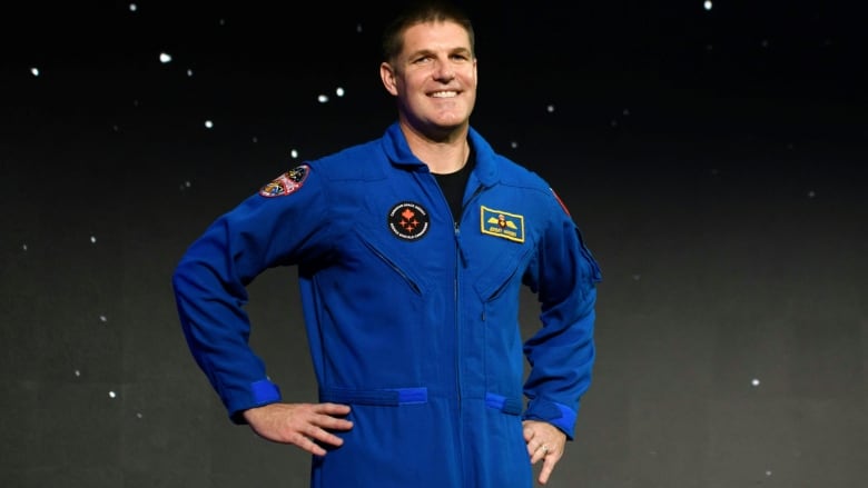 A man wearing a blue space jumpsuit smiles, with his hands on his hips, in front of a display screen showing a starry night.