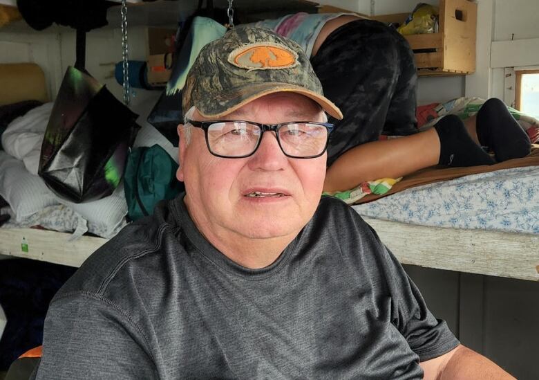 A man in glasses and a ballcap sits in a chair.