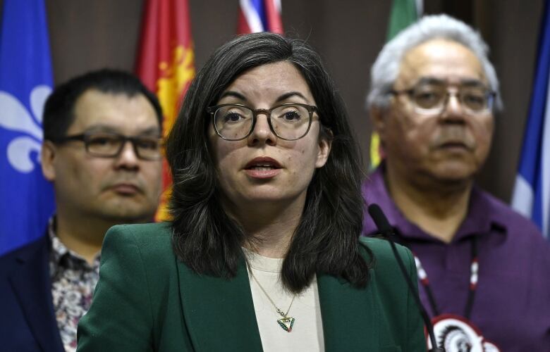 A woman with glasses and a green blazer speaks into a microphone at a podium. Two people stand behind her.