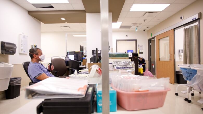 A man sits on one side of a glass panel at a desk, he's wearing medical scrubs and a COVDI-19 face mask. 