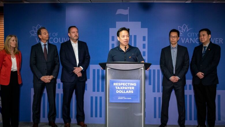 A man, flanked by four other people, speaks from a podium with a sign on it that says, Respecting Taxpayer Dollars.