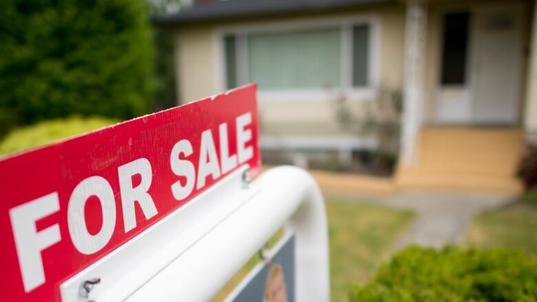 A for sale sign is in focus staked into a lawn with what appears to be a single-family home blurred in the background. 