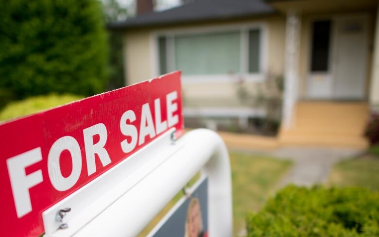 A for sale sign is in focus staked into a lawn with what appears to be a single-family home blurred in the background. 