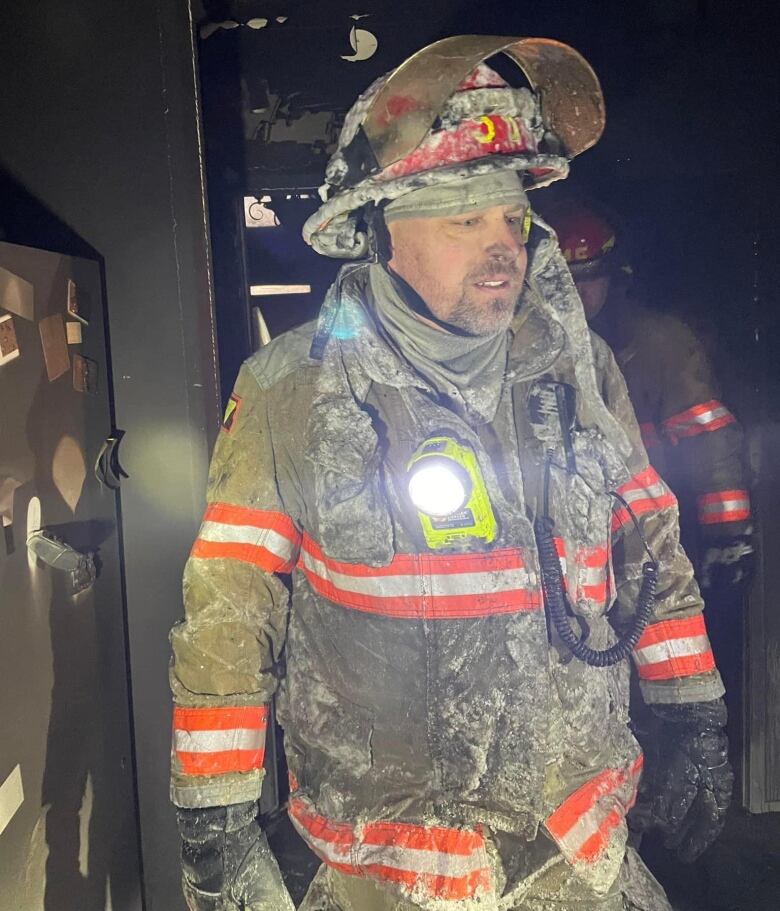 A man stands in a house kitchen in firefighting gear that's covered in debris. His nose is blackened with soot.