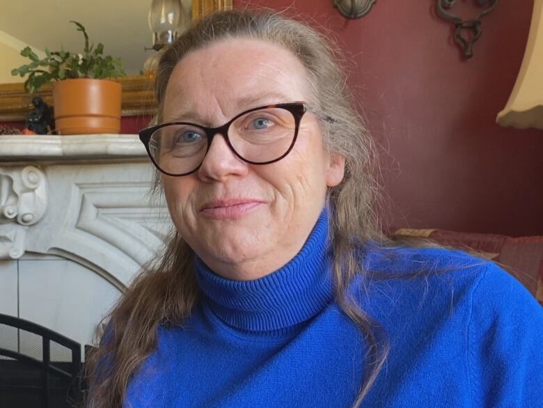 A woman with long brown hair and glasses wearing sits in front of a fireplace. She is wearing a blue top.