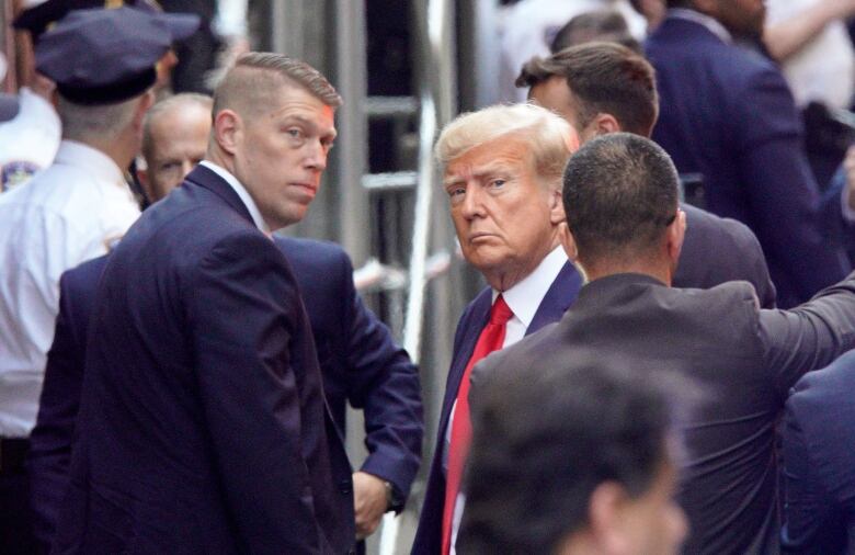 Donald Trump, with a blank expression, surrounded by men in suits and by police officers.