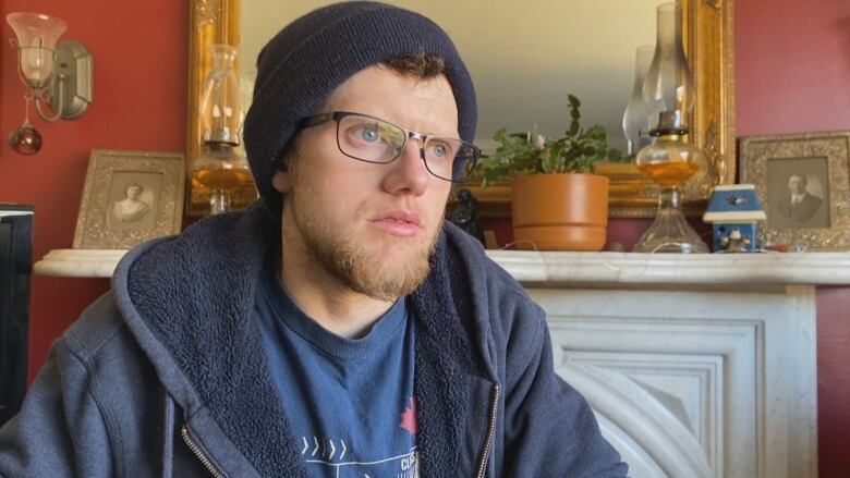 A man wears a tuque and glasses with a navy zip-up hoodie and t-shirt sits in front of a fire place.