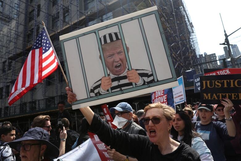 Cartoon of Trump behind bars, on a protest sign being held up by a woman in sunglasses who is also carrying an American flag. 