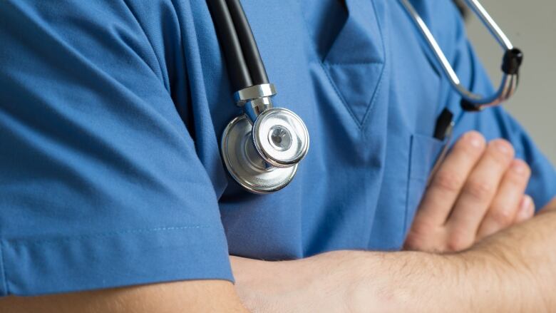 A close-up of a doctor in blue scrubs with a stethoscope around their neck and their arms crossed.