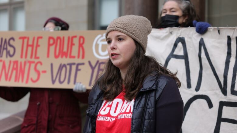 A young woman in a toque and t-shirt reading 'ACORN' stands in front of signs held up by demonstrators.