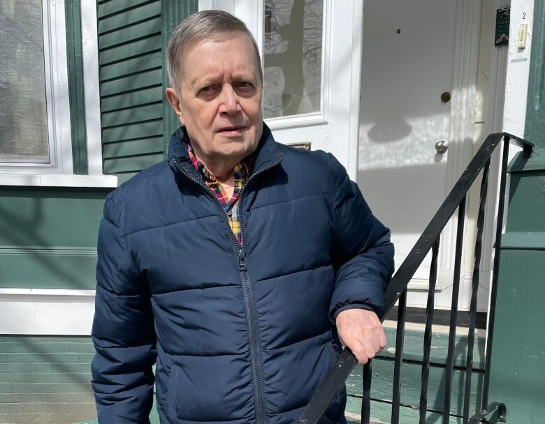 A man wearing a navy blue coat standing in front of a blue building while holding onto the rail by the steps to the doorway.