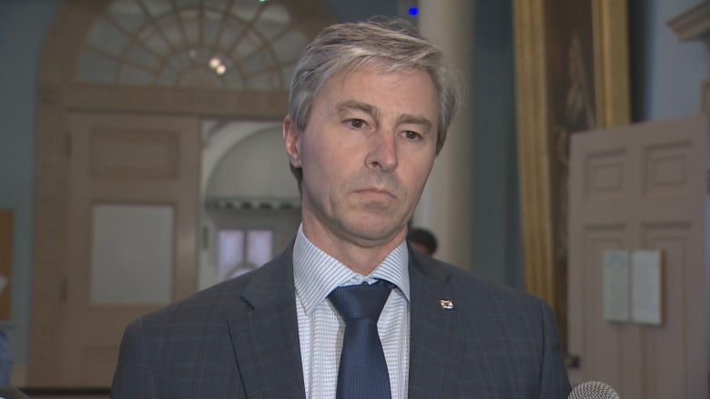 A man with grey hair wears a dark grey blazer with a striped shirt and dark blue tie. He is standing near an entranceway. 