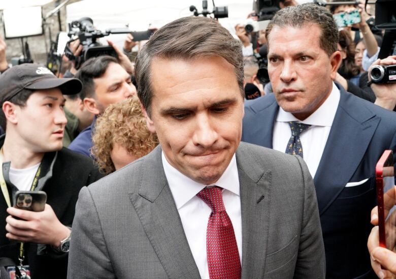 Man in grey suit and red tie, surrounded by reporters