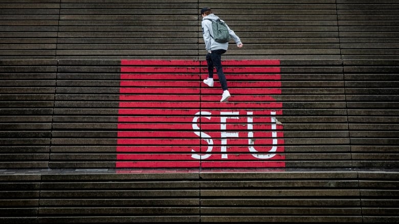 A person in a hoodie walks over steps that have 'SFU' and a red logo painted over in a gradient.