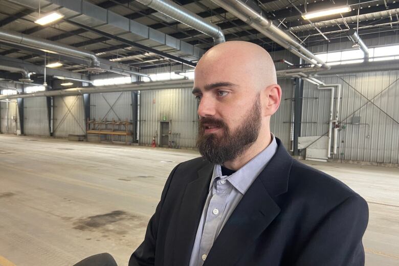 A man with a beard stands inside a big open warehouse building.