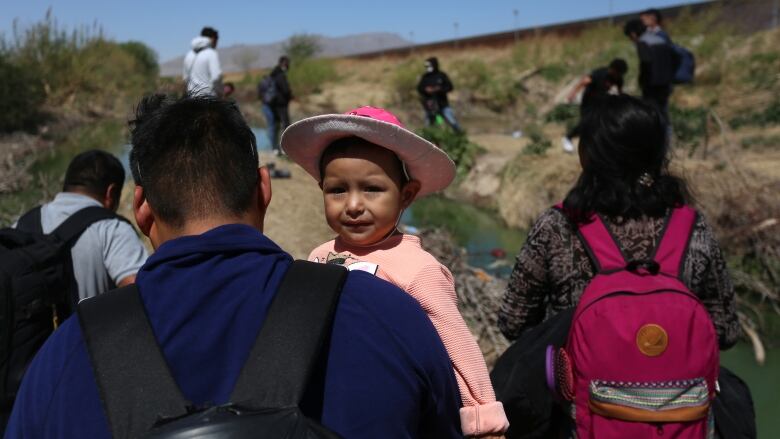 A small child, wearing a hat, is carried by an adult wearing a backpack among a group of other people crossing in hot, desert like environment. There is a fence in the distance.