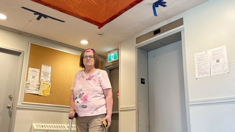 An older woman stands with a cane in her apartment lobby, where there's a large orange tarp covering a hole caused by a water leak.