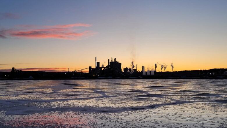 A mill with smokestacks silhouetted against a vibrant sunset is seen in the distance across a frigid body of water.