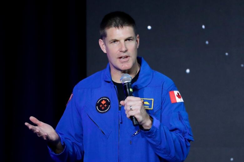 The Canadian astronaut is on stage speaking into a microphone in his hand while wearing NASA astronaut jumpsuit.