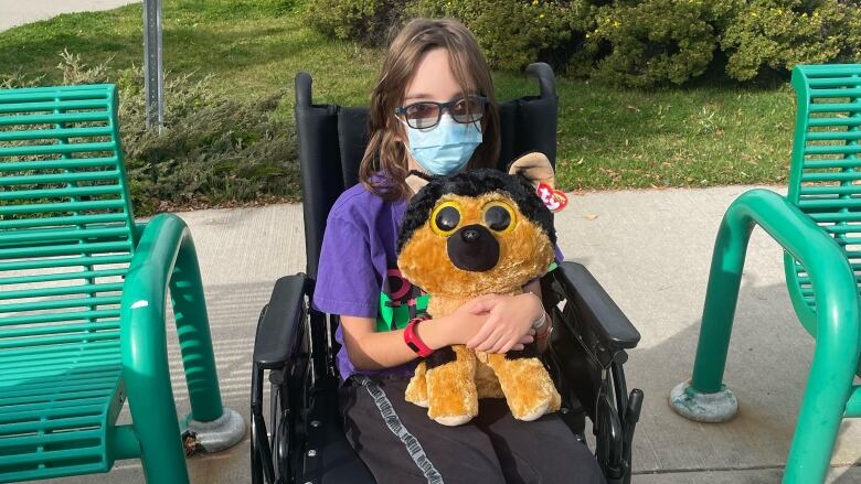 Keegan is sitting in a wheelchair outside Alberta Children's Hospital, wearing a mask, sunglasses and holding a teddy bear.