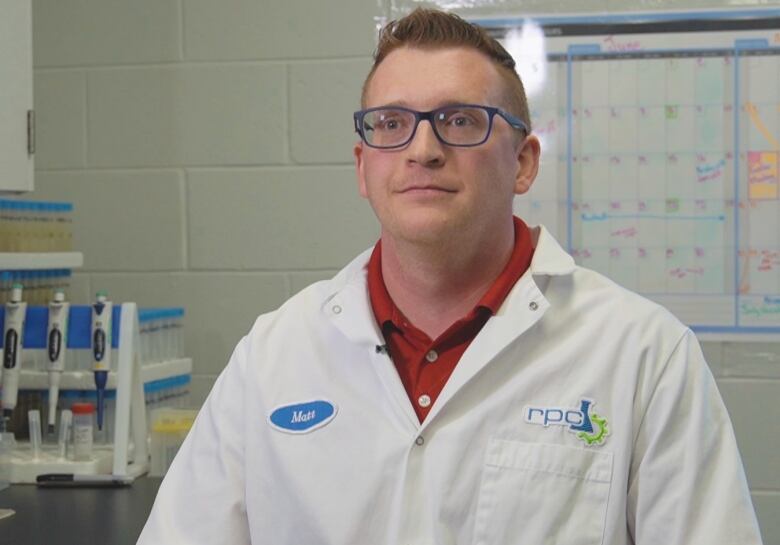 A man wearing a lab coat and glasses sitting in a lab with white brick walls.