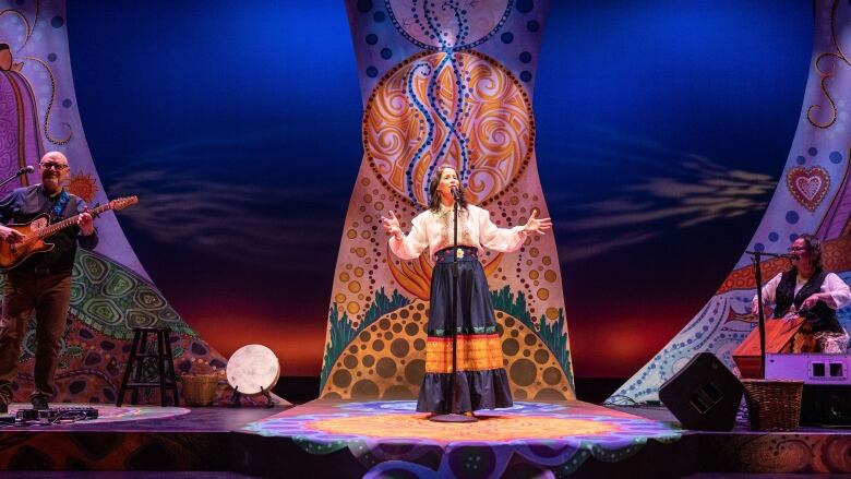 A woman in a white shirt and long blue skirt, stands in the middle of a stage with her arms stretched wide while singing into a microphone. Behind her, the stage is decorated in an intricate pattern of circles and swirls. 