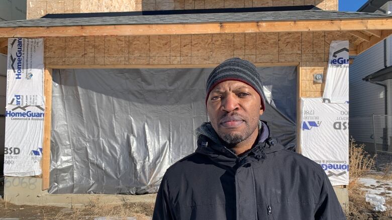 A man wearing a black jacket and hat stands in front of an unfinished house.