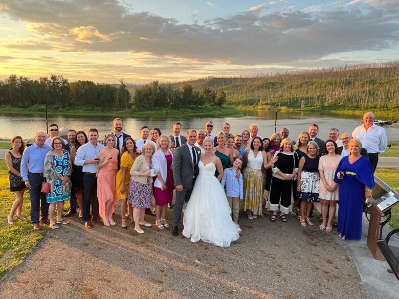 A wedding party stands in front of a body of water with a fire-damaged forest behind.