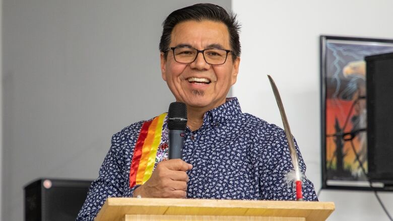 A man has short black hair, eyeglasses and wears a floral shirt with red, orange and yellow stripes down the right side. He stands at a podium speaking into a microphone held in his hand.