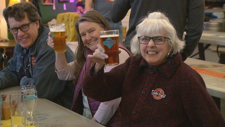 Two women and one man hold beers up to the camera in a 