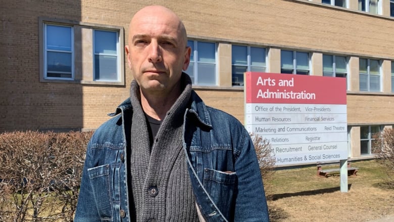 A bald man wearing a layered sweater and jean jacket stands outside a building. A sign which reads 