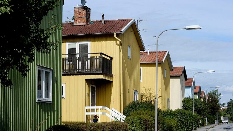 A street is lined with a green hedge and two-storey green, yellow, blue and white homes with red roofs.