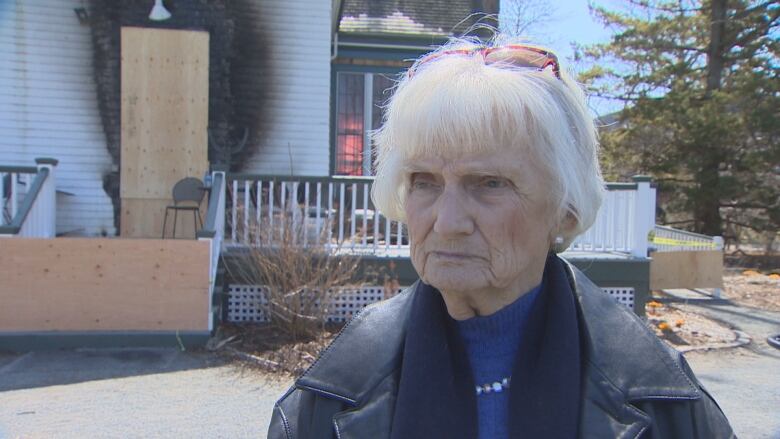 A woman with short grey hair wearing a black leather jacket stands in front of a burnt house-like structure.