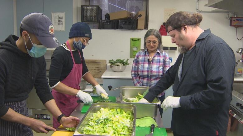 Four people stand in a kitchen, three of them chopping leeks into a metal container.