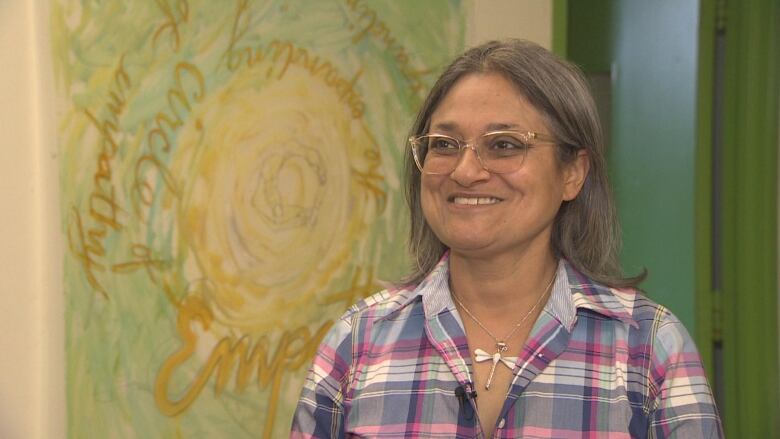 A woman with grey hair and glasses, wearing a pink and blue plaid shirt, smiles at the camera. 