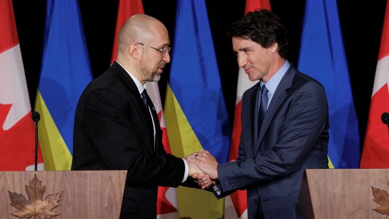 While smiling warmly, a man clasps the hands of another man at a podium in a Toronto hotel.