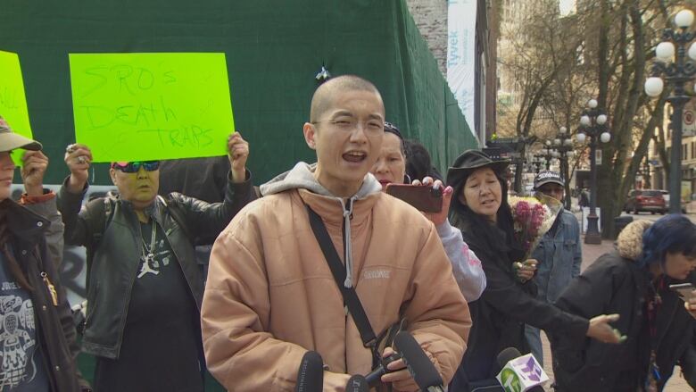 A bald man speaks at a rally, with a sign behind him reading 'SROs death traps'.