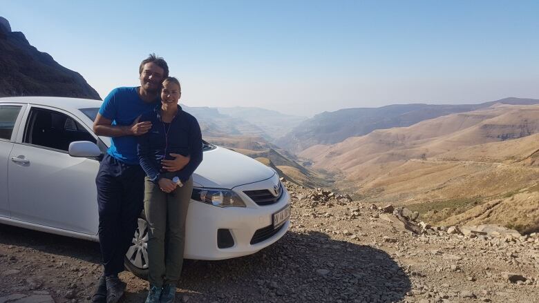 A man and woman stand in front of a white car on top of a mountain.