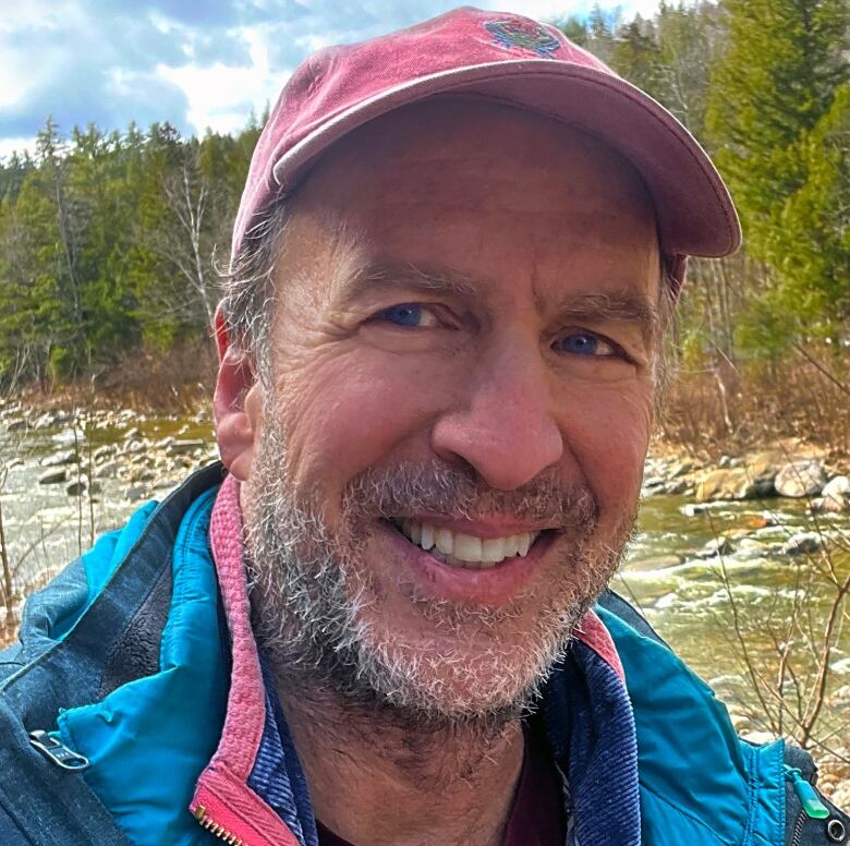 A man outside smiling. He is wearing a blue jacket and red baseball cap.