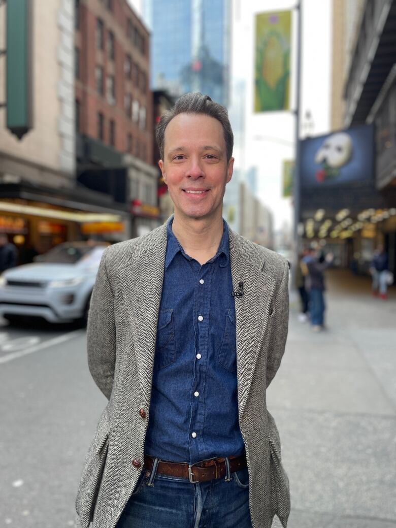 A man wearing a sports jacket and jeans stands in front of a theatre house.