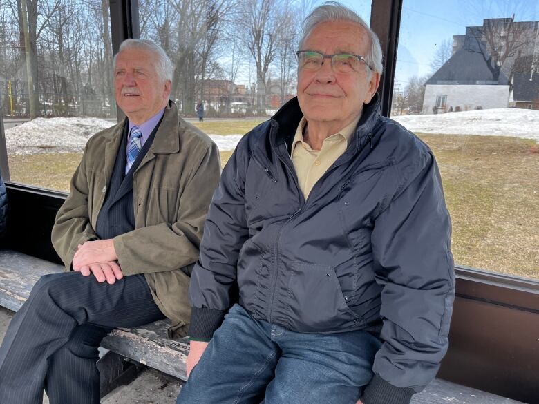 Two grey-haired men sit on a bench and look into the camera.