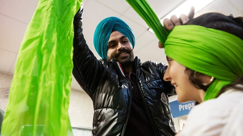 Man wearing black puffer jacket with a mustache and blue turban ties a green turban on a beaming person.