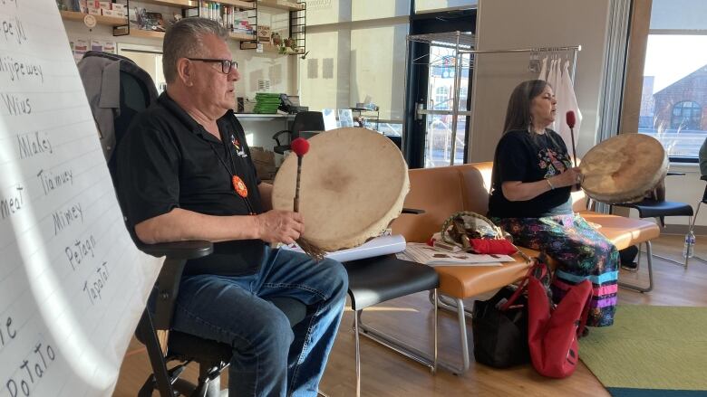 A man sits to the left of a woman in what looks like a classroom. They are both holding traditional Mi'kmaq drums with drum sticks in the air.