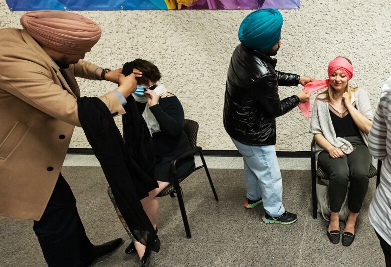 Three people sit in chairs while three men, two of whom wear turbans, tie turbans around the heads of those sitting.