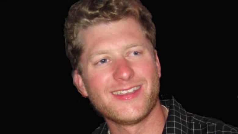 A man with wavy blonde hair wears a dark checkered shirt and smiles for a photo.